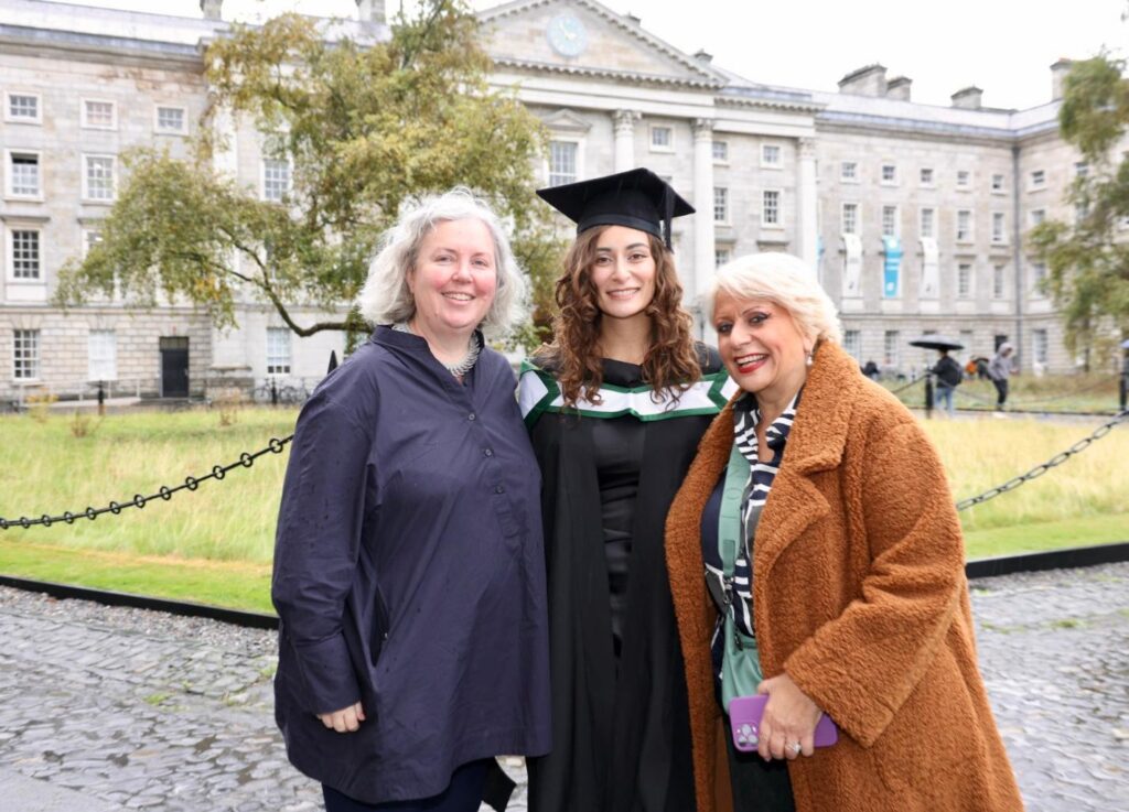 Three women stand together, smiling at the camera. One of them is wearing a graduation cap and gown.