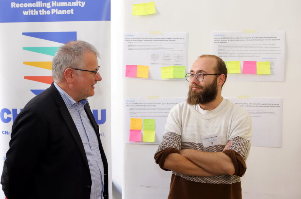 Two participants at the libraries meet-up discussing with the CHARM-EU rollup on the back and a panel with notes and post-its.