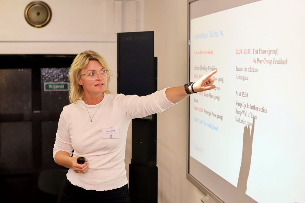 Prof. Dr. Simone Roth from Hochschule Ruhr West is explaining the agenda of the Design Thinking Day pointing at the screen. 