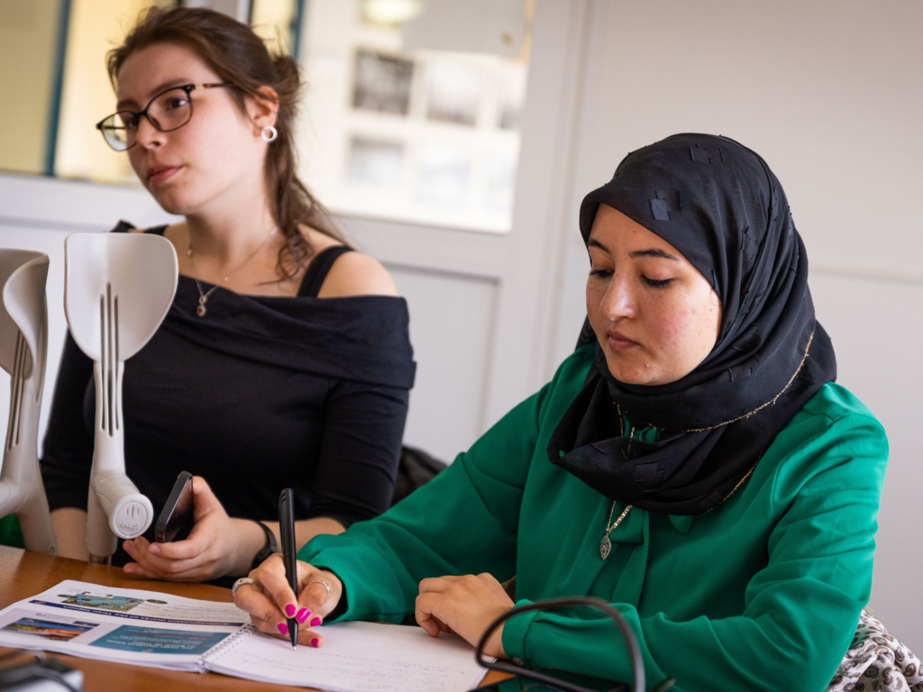 Two women in a class. The one on the left is listening and the other on the right is writing in a notebook