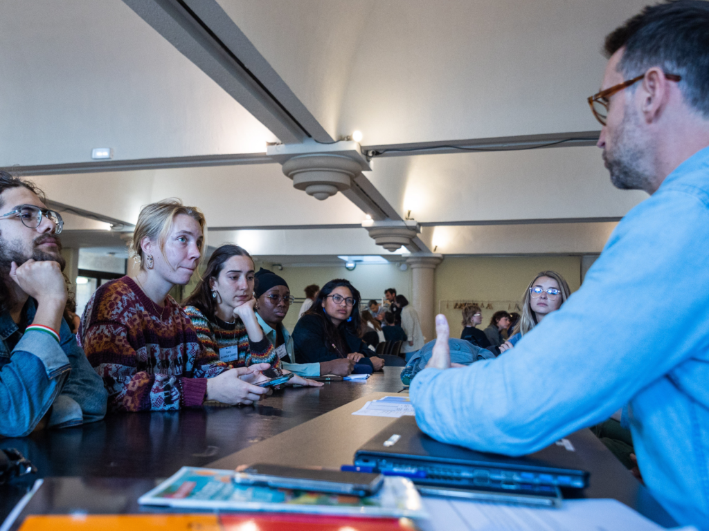 A group of people of different nationalities, sitting around a large table and listening to a teacher.