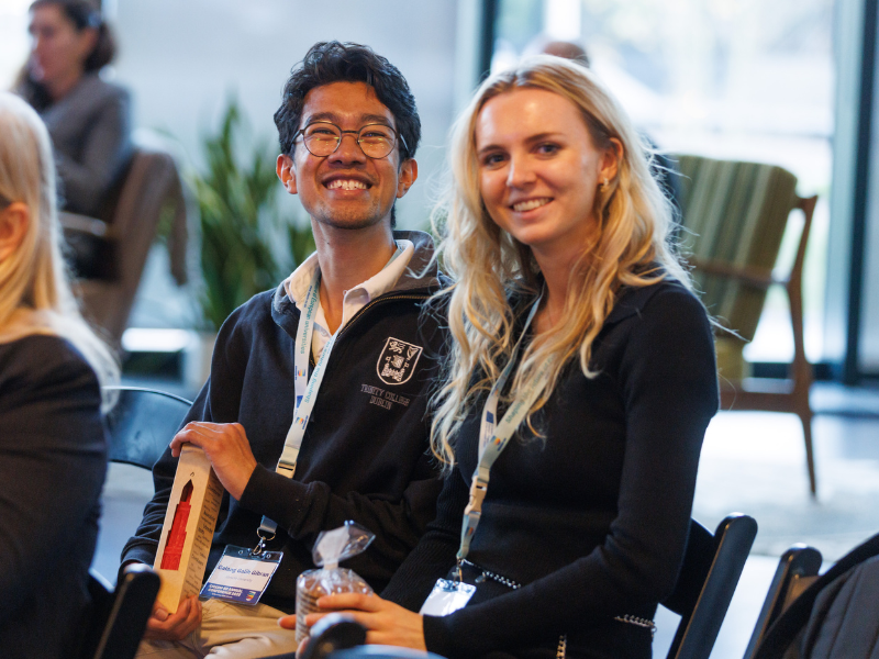 Two people are seated next to each other at an event, wearing lanyards with name badges. One person is holding a rectangular award or gift box, while the other is holding a small wrapped item. They are seated among other attendees in a well-lit indoor space with chairs and plants in the background.