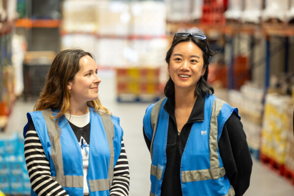 Two young women stand together in a. warehouse. They are both wearing blue high-vis vests and are smiling
