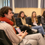 A young man with curly hair and a red scarf sits in a lecture hall chair, gesturing with his hands as he contributes to a group discussion. Three other students are visible in the background, listening attentively.