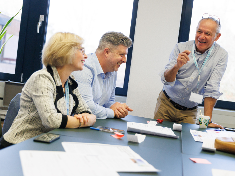 Three people sit and stand around a meeting table, engaged in discussion. Papers, notebooks, pens, and a smartphone are spread across the table, and one person is gesturing while speaking. The setting appears to be a bright office or meeting room with large windows in the background.