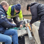 Three individuals working together on a small metal boat, handling ropes and equipment. One person kneels while adjusting a cylindrical device attached to the boat. A brick wall and canal water are visible in the background.