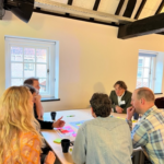 Several people seated around a table in a bright room with wooden beams on the ceiling. They are engaged in discussion, with papers, colorful sticky notes, and mugs spread across the table. Two windows provide natural light.