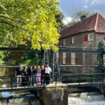A group of people standing on a small bridge above flowing water, surrounded by lush greenery. Behind them is a historic brick building with a red‑tiled roof and red window shutters, next to a calm canal.