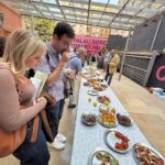 An outdoor tasting or exhibition area with a long table covered in small plates of various foods. People stand along the table examining and serving themselves from the displayed dishes. A large sign in the background reads “PALAU ROBERT.”