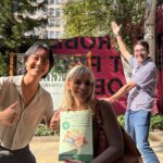 Three people standing outdoors in a sunny area with trees and buildings in the background. One person in front is holding a brochure, while another gives a thumbs‑up gesture. A third person in the background raises both arms enthusiastically. A large pink sign with bold lettering is partially visible behind them.