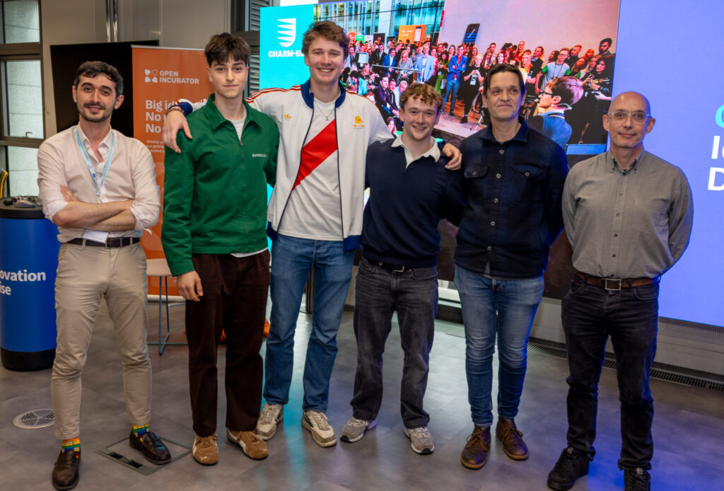 A group of people stand together in front of a large screen. Some of them have their arms around each other, they are all smiling at the camera.