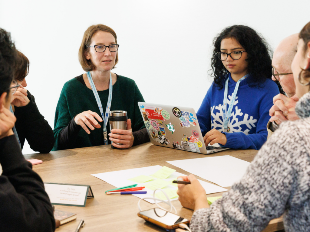 A diverse group of five people sits around a wooden conference table in a bright, white room, engaged in a collaborative meeting. A woman with glasses and a green sweater gestures while speaking, holding a reusable coffee cup. To her right, a young woman with curly hair and a blue snowflake sweater types on a laptop covered in colorful stickers. In the foreground, hands are visible holding a pen over yellow sticky notes and blank paper, alongside a smartphone and pens. Everyone is wearing blue lanyards, suggesting a professional or academic workshop setting.