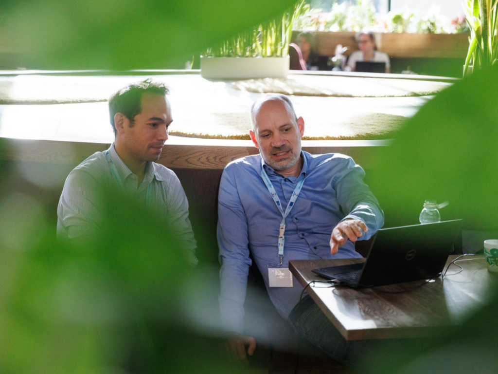 Two men sitting side-by-side in a modern, sunlit indoor space collaborate on a laptop. The man on the right, wearing a blue button-down shirt and an event lanyard, gestures toward the screen while speaking. The man on the left, wearing a light-striped button-down shirt, listens intently while looking at the laptop. Green foliage softly blurs the foreground, framing the shot.