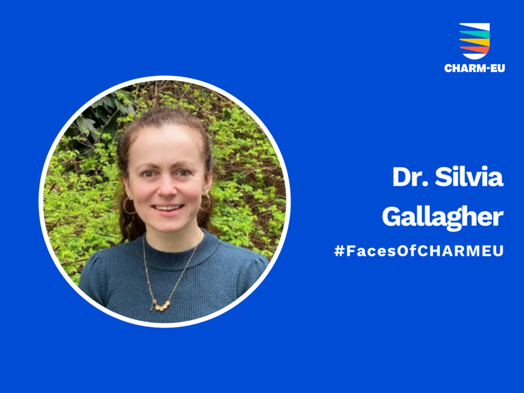 A professional graphic on a solid blue background featuring a circular headshot of Dr. Silvia Gallagher on the left. She is smiling, with dark hair tied back, wearing a blue sweater and a gold necklace, positioned in front of green foliage. To the right of the photo, the white text reads "Dr. Silvia Gallagher" followed by the hashtag "#FacesOfCHARMEU." The top right corner contains the CHARM-EU logo, which consists of a stylized shield with colorful horizontal stripes.