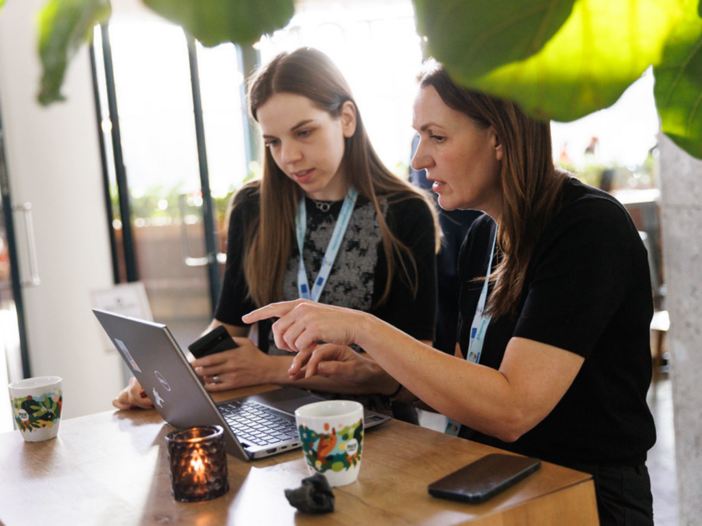 Two colleagues sitting at a wooden table in a bright, modern indoor space work together on a laptop. The person on the right, wearing a black shirt, points at the laptop screen while explaining something. The person on the left, holding a smartphone, looks closely at the screen. Both are wearing light blue event lanyards. On the table are two patterned coffee mugs, a smartphone, and a small lit candle holder, with green leaves softly blurring the top right foreground.