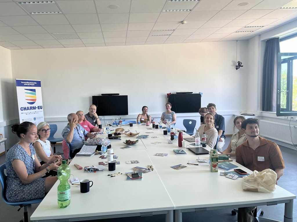 Participants seated around a table during a CHARM‑EU workshop, listening to a presentation in a meeting room.