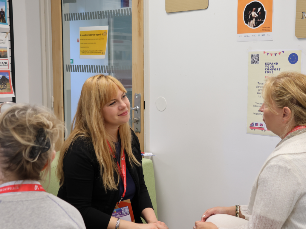 An indoor scene featuring three women engaged in a conversation. In the center, a blonde woman with long hair and a black blazer sits facing right, smiling warmly and listening intently. She wears a red lanyard with ID badges around her neck. To her right, another blonde woman, viewed from a side profile and wearing a light-colored cardigan, sits facing her. In the foreground on the left, the back of a third woman's head is visible, showing blonde hair tied up and a red lanyard around her neck. The background shows a white wall decorated with posters—including one with the text "EXPAND YOUR COMFORT ZONE"—and a glass door with a wooden frame featuring a yellow safety sign.