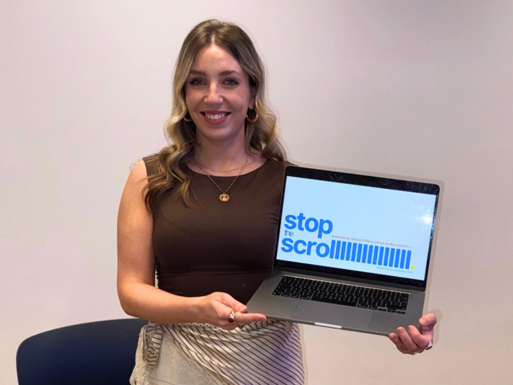 A smiling woman with wavy blonde hair stands against a plain light wall, holding an open laptop towards the camera. She is wearing a brown sleeveless top, necklaces, and a striped wrap around her waist. The laptop screen displays a graphic with the bold blue text "stop the scroll", followed by a series of vertical blue lines and smaller, lighter text that reads "translating research for a social media audience."