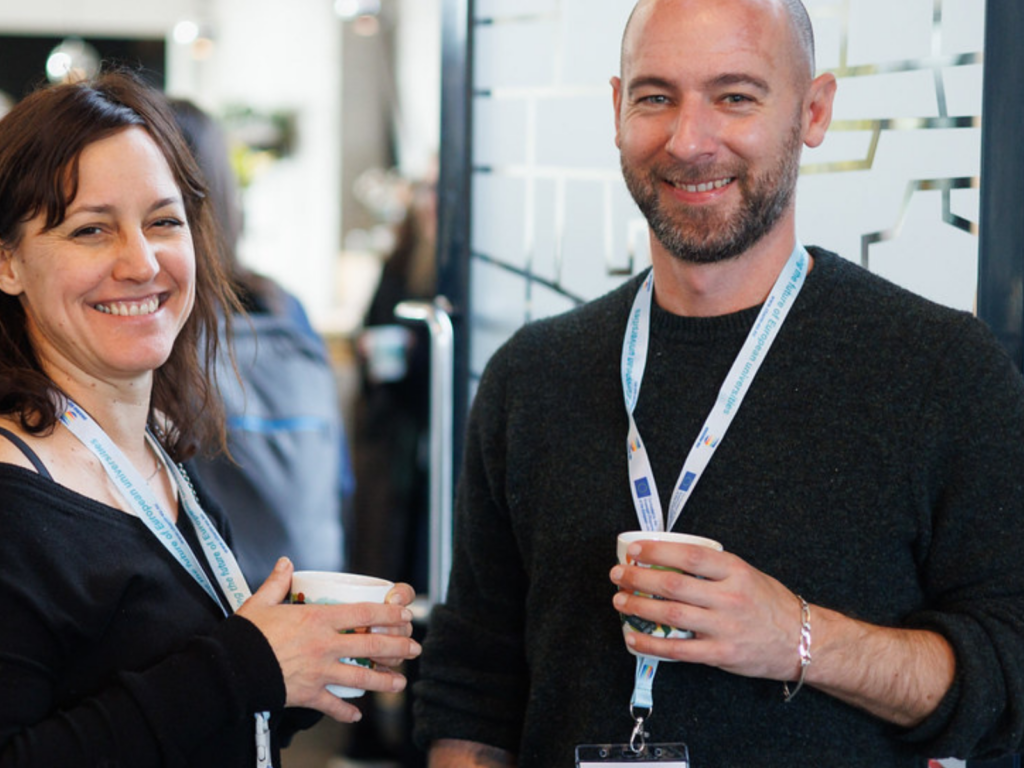 Here is an alternative text description for the image "Peer Coaching - Thrive Together.jpg": **Alt Text:** A man and a woman stand close together smiling at the camera in an indoor setting. Both are wearing lanyards and holding white mugs. The woman on the left has dark brown hair and is wearing a black shirt, while the bald man on the right has a short beard and is wearing a dark grey sweater. The background is slightly blurred with other people visible.