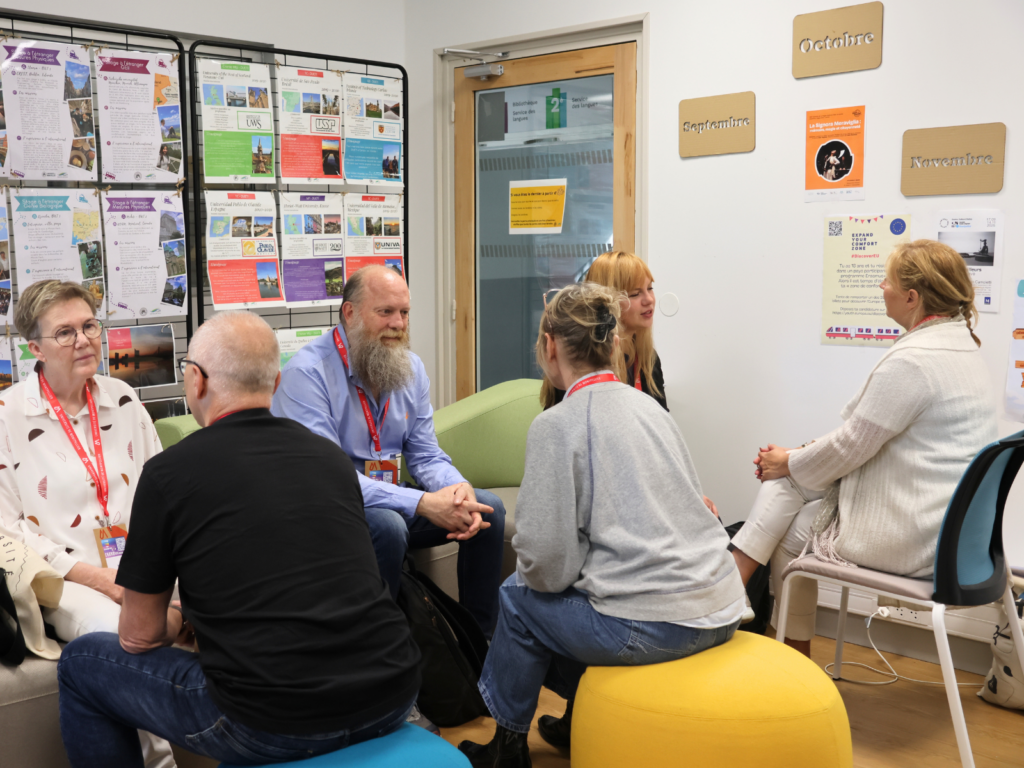 A group of six people sit in a circle in an office or classroom setting, engaged in conversation. On the left, an older woman with glasses and a patterned blouse sits opposite a man with a long gray beard and a blue button-down shirt. Two individuals sit with their backs to the camera in the foreground—one on a blue pouf and another on a yellow pouf. On the right, two blonde women are seated, one in a white cardigan facing the group. The background features a glass-paned door, a wall with signs for "Septembre," "Octobre," and "Novembre," and a display rack filled with colorful informational brochures.