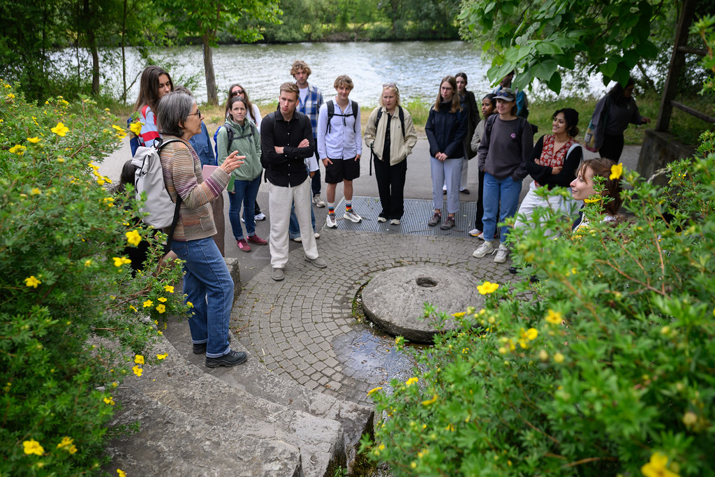Students gathered outdoors by a riverside, listening to a guide during an educational field visit.