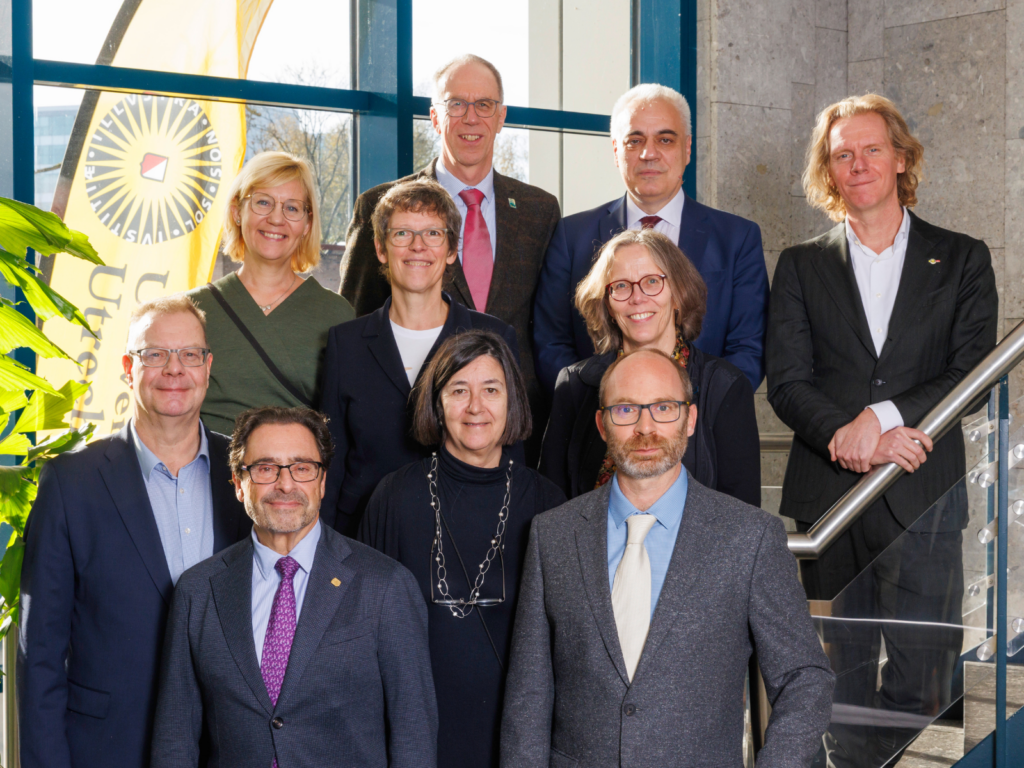 Group portrait of nine Rectors and Vice-rectors from the CHARM-EU partner universities, standing on a staircase indoors next to a large window and an Utrecht University banner. The individuals—five women and four men—are dressed in business casual and professional attire, arranged in three rows, and smiling at the camera.