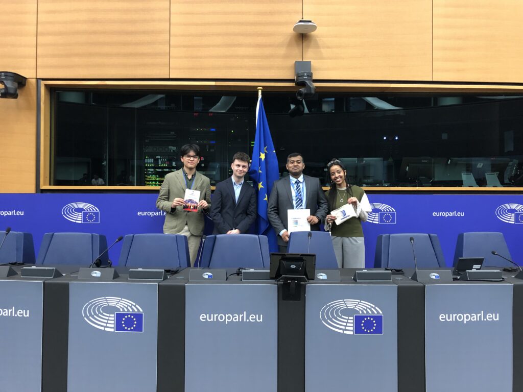 Large group photo of participants gathered inside the European Parliament chamber beneath EU flags.