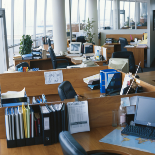 A wide shot of a modern, open-plan office. Numerous wooden desks with low dividers are neatly arranged, creating designated workspaces. The office is empty of people, but the desks are equipped with desktop computers (including a mix of older CRT-style and flatter monitors), landline phones, and various office supplies like ring binders, papers, and small potted plants. Tall white pillars and floor-to-ceiling windows run along the left wall, letting in bright daylight. The scene is clean, organized, and ready for a workday.