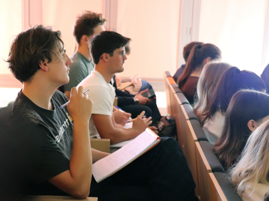 A row of students seated in tiered wooden desks, taking notes in an auditorium or classroom. In the foreground, a young man with curly brown hair wearing a black shirt and glasses holds a pen and an open notebook, looking towards the left, outside the frame. Further back, another student in a white shirt is also looking attentively, while a third is looking down at a mobile device. The background is brightly lit by a window, creating a lens flare and obscuring the faces of others. The focus is primarily on the foreground student and his open notebook.
