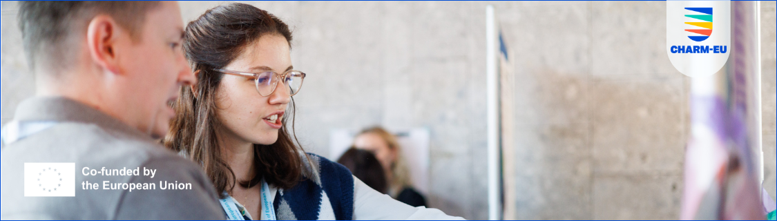 A candid medium shot of a young woman with long brown hair and round glasses, smiling as she speaks with a man whose back is partially to the camera. They appear to be in a professional or academic setting, possibly viewing a poster or display just out of frame. The background is a bright, neutral wall. The top right corner of the image contains a white speech bubble graphic featuring a multi-colored (green, blue, orange) emblem of horizontal lines forming a stack and the text "CHARM-EU." The bottom left corner has a white square logo with the European Union flag (circle of yellow stars on a blue field) and the text "Co-funded by the European Union." The overall impression is one of collaboration and EU-supported activity.