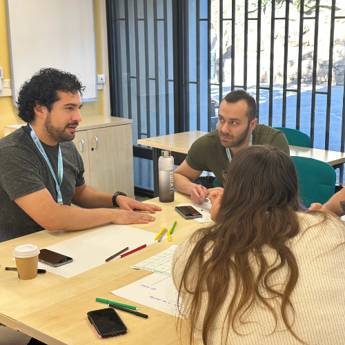 The image shows a group of three people, two men, and one woman, sitting around a table with large sheets of paper, ready for work. They look at each other, presumably discussing the project. On the table, you can also see colored pens, a coffee mug, smartphones, and a water bottle. In the background, there is a window, and to the left, there is a cabinet with a small whiteboard above it.