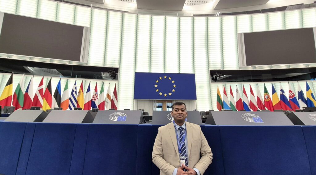 Participant posing in the European Parliament chamber with EU and member state flags in the background.