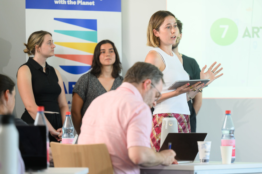 Participant presenting to a group in a classroom, with a CHARM‑EU banner visible in the background.