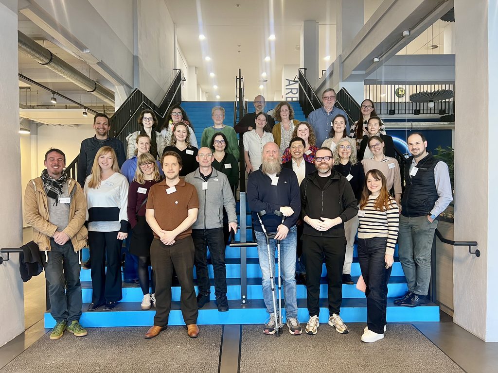 Group photo of participants standing on a staircase inside a modern university building during a CHARM‑EU meeting.