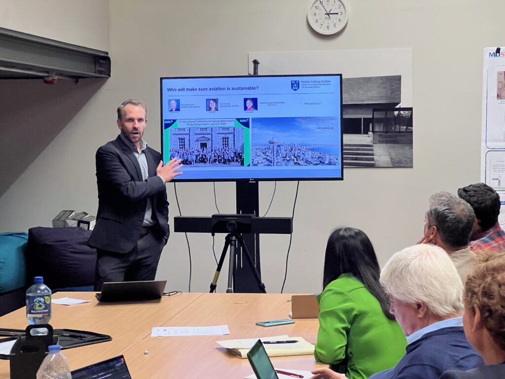 Participant presenting research findings to a group during a meeting, with a presentation screen and research posters visible.
