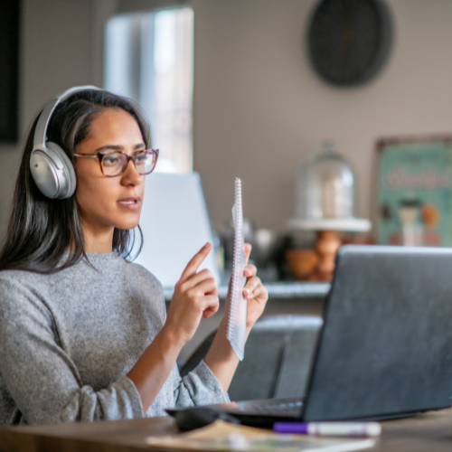Person wearing headphones participating in an online language class, speaking to a laptop while taking notes in a notebook.