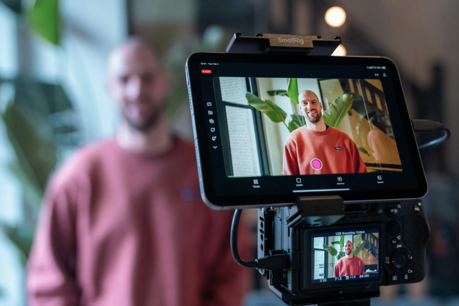 Person being filmed on a professional camera setup, with the camera screen showing a live recording in an indoor setting with plants in the background.