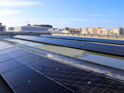 A wide view of several rows of solar panels installed on a flat rooftop, arranged in parallel lines. In the background, multiple low-rise buildings are visible under a clear blue sky.