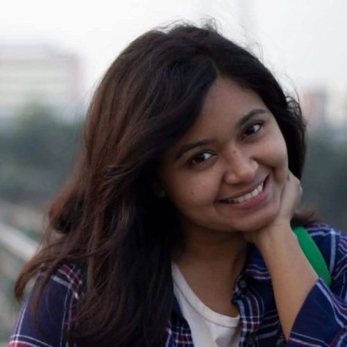 A close-up portrait photograph shows a young woman with shoulder-length dark brown hair, looking directly at the camera with a cheerful, wide smile that shows her teeth. She has her hand, with her fingers slightly curled, resting against her chin. She wears a navy blue plaid button-down shirt over a white t-shirt and has a green bag strap visible over her left shoulder. The background is an out-of-focus cityscape with buildings under an overcast sky.