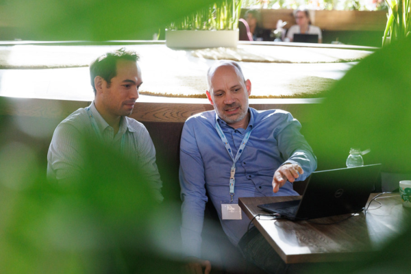 Two men sitting side-by-side in a modern, sunlit indoor space collaborate on a laptop. The man on the right, wearing a blue button-down shirt and an event lanyard, gestures toward the screen while speaking. The man on the left, wearing a light-striped button-down shirt, listens intently while looking at the laptop. Green foliage softly blurs the foreground, framing the shot.