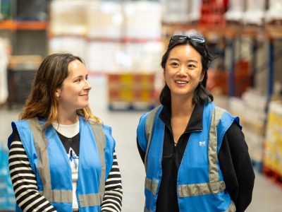 Two students stand in a warehouse. They are wearing blue high-vis vests