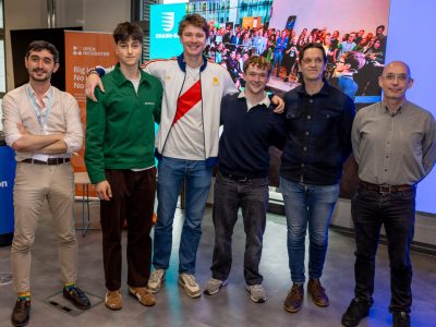 A group of people stand together in front of a large screen. Some of them have their arms around each other, they are all smiling at the camera.