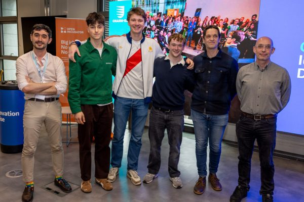 A group of people stand together in front of a large screen. Some of them have their arms around each other, they are all smiling at the camera.