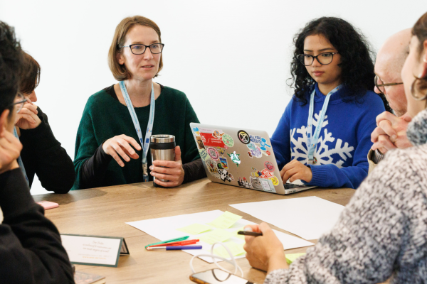 A diverse group of five people sits around a wooden conference table in a bright, white room, engaged in a collaborative meeting. A woman with glasses and a green sweater gestures while speaking, holding a reusable coffee cup. To her right, a young woman with curly hair and a blue snowflake sweater types on a laptop covered in colorful stickers. In the foreground, hands are visible holding a pen over yellow sticky notes and blank paper, alongside a smartphone and pens. Everyone is wearing blue lanyards, suggesting a professional or academic workshop setting.
