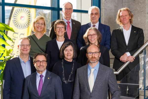 Group portrait of nine Rectors and Vice-rectors from the CHARM-EU partner universities, standing on a staircase indoors next to a large window and an Utrecht University banner. The individuals—five women and four men—are dressed in business casual and professional attire, arranged in three rows, and smiling at the camera.