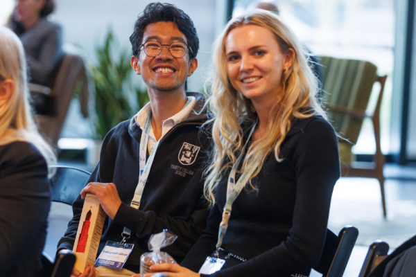 Two people are seated next to each other at an event, wearing lanyards with name badges. One person is holding a rectangular award or gift box, while the other is holding a small wrapped item. They are seated among other attendees in a well-lit indoor space with chairs and plants in the background.