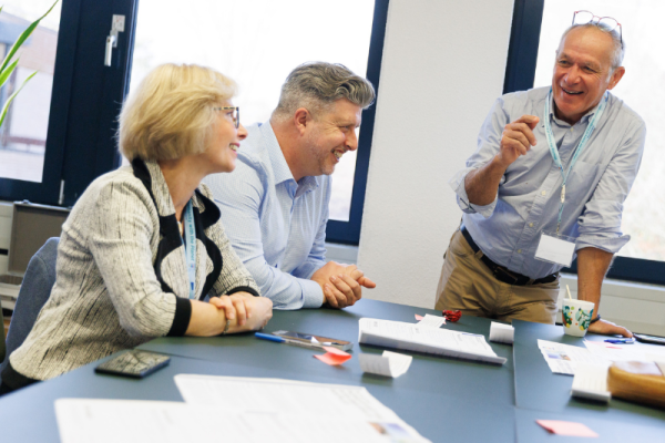 Three people sit and stand around a meeting table, engaged in discussion. Papers, notebooks, pens, and a smartphone are spread across the table, and one person is gesturing while speaking. The setting appears to be a bright office or meeting room with large windows in the background.
