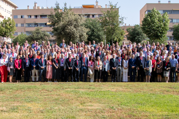 A large group of people pose for a group photo in an outdoor park setting with trees and apartment buildings in the background.