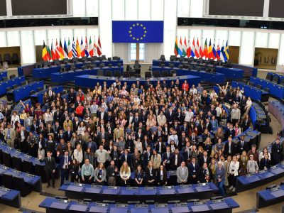 Large group photo of participants gathered inside the European Parliament chamber beneath EU flags.