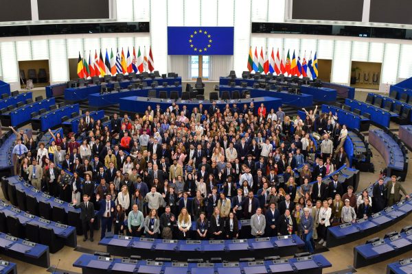 Large group photo of participants gathered inside the European Parliament chamber beneath EU flags.